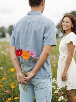 One partner hiding a bunch of picked flowers behind their back in a flower field